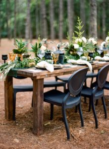 farmtable with black chairs and emerald and gold accents nestled in the woods for a woodsy wedding or holiday party
